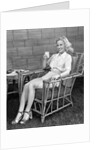 1940s blonde woman in bamboo chair holding up glass of milk smiling wearing white shorts looking at camera by Anonymous