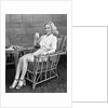1940s blonde woman in bamboo chair holding up glass of milk smiling wearing white shorts looking at camera by Anonymous
