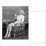 1940s blonde woman in bamboo chair holding up glass of milk smiling wearing white shorts looking at camera by Anonymous