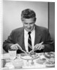 1950s smiling man in suit and tie sitting at table holding knife and fork eating dinner by Anonymous