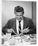 1950s smiling man in suit and tie sitting at table holding knife and fork eating dinner by Anonymous