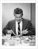 1950s smiling man in suit and tie sitting at table holding knife and fork eating dinner by Anonymous