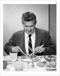 1950s smiling man in suit and tie sitting at table holding knife and fork eating dinner by Anonymous