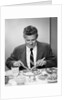 1950s smiling man in suit and tie sitting at table holding knife and fork eating dinner by Anonymous