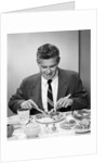 1950s smiling man in suit and tie sitting at table holding knife and fork eating dinner by Anonymous