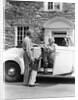 1940s senior retired couple loading golf clubs into white convertible car in front stone house by Anonymous
