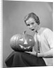1950s woman wearing sweater holding carved pumpkin jack-o-lantern on her lap by Anonymous