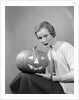 1950s woman wearing sweater holding carved pumpkin jack-o-lantern on her lap by Anonymous