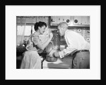 1950s housewife in kitchen having husband taste food on stove by Anonymous