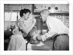 1950s housewife in kitchen having husband taste food on stove by Anonymous