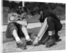 1950s teen boy helping girl put on metal roller skates sitting on sidewalk by Anonymous