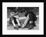 1950s teen boy helping girl put on metal roller skates sitting on sidewalk by Anonymous