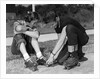1950s teen boy helping girl put on metal roller skates sitting on sidewalk by Anonymous
