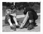 1950s teen boy helping girl put on metal roller skates sitting on sidewalk by Anonymous