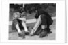 1950s teen boy helping girl put on metal roller skates sitting on sidewalk by Anonymous