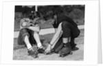 1950s teen boy helping girl put on metal roller skates sitting on sidewalk by Anonymous