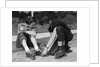 1950s teen boy helping girl put on metal roller skates sitting on sidewalk by Anonymous