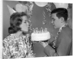 1960s teenage girl blowing out candles on birthday cake held by boy by Anonymous