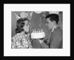 1960s teenage girl blowing out candles on birthday cake held by boy by Anonymous