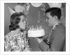 1960s teenage girl blowing out candles on birthday cake held by boy by Anonymous