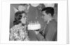 1960s teenage girl blowing out candles on birthday cake held by boy by Anonymous