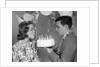 1960s teenage girl blowing out candles on birthday cake held by boy by Anonymous