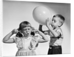 1950s little boy blowing up big balloon little girl with fingers in ears eyes closed by Anonymous