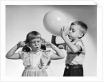 1950s little boy blowing up big balloon little girl with fingers in ears eyes closed by Anonymous