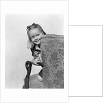 1940s little girl holding doll peeking around side of chair by Anonymous