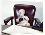 1960s chubby baby sitting in leather chair at office desk holding telephone by Anonymous