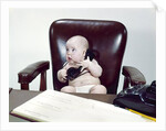1960s chubby baby sitting in leather chair at office desk holding telephone by Anonymous