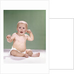 1960s baby wearing diaper sitting on pink blanket mouth open hand touching ear by Anonymous