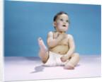 1960s baby wearing diaper sitting looking up and kicking one leg up by Anonymous