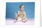 1960s baby wearing diaper sitting looking up and kicking one leg up by Anonymous