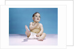 1960s baby wearing diaper sitting looking up and kicking one leg up by Anonymous
