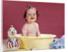 1960s smiling brunette baby sitting in yellow plastic bath tub with toy clown looking at camera by Anonymous