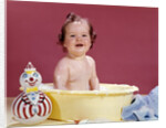 1960s smiling brunette baby sitting in yellow plastic bath tub with toy clown looking at camera by Anonymous