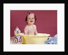 1960s smiling brunette baby sitting in yellow plastic bath tub with toy clown looking at camera by Anonymous