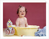 1960s smiling brunette baby sitting in yellow plastic bath tub with toy clown looking at camera by Anonymous