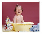 1960s smiling brunette baby sitting in yellow plastic bath tub with toy clown looking at camera by Anonymous