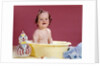 1960s smiling brunette baby sitting in yellow plastic bath tub with toy clown looking at camera by Anonymous
