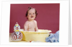 1960s smiling brunette baby sitting in yellow plastic bath tub with toy clown looking at camera by Anonymous