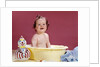 1960s smiling brunette baby sitting in yellow plastic bath tub with toy clown looking at camera by Anonymous