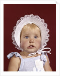 1960s portrait baby girl wearing bonnet and blue gingham dress showing two front teeth looking at camera by Anonymous