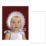 1960s portrait baby girl wearing bonnet and blue gingham dress showing two front teeth looking at camera by Anonymous