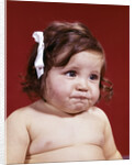 1960s portrait of chubby unhappy baby girl with white ribbon bow in messy hair by Anonymous