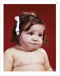 1960s portrait of chubby unhappy baby girl with white ribbon bow in messy hair by Anonymous
