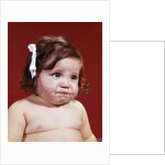 1960s portrait of chubby unhappy baby girl with white ribbon bow in messy hair by Anonymous