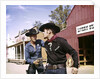 1960s cowboy sheriff with badge draws gun arrests gunfighter in front of livery stable by Anonymous