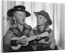 1950s 2 juvenile boys in cowboy hat & shirts playing ukulele & singing mouth open wide by Anonymous